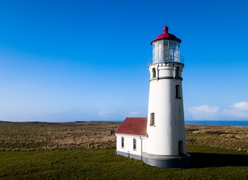 A white lighthouse with a red roof stands on a grassy landscape under a clear blue sky.