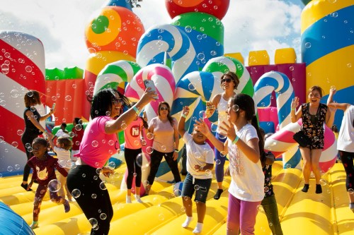 A lively scene of children and adults playing on a colorful inflatable structure, surrounded by bubbles and beach balls.