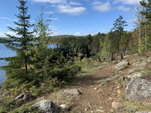 A scenic view of a trail by a lake, surrounded by trees and rocky terrain under a blue sky with scattered clouds.