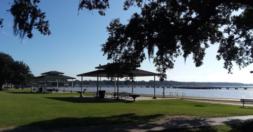 A serene lakeside park with pavilions, green grass, and a clear blue sky reflecting on the water.