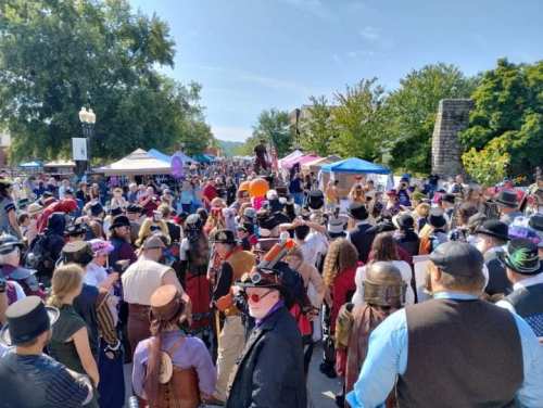 A lively street festival with a crowd in costumes, tents, and trees under a clear blue sky.