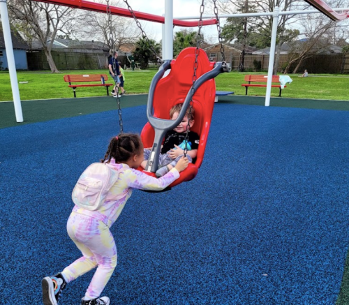 A child in a red swing is being pushed by another child on a playground with blue flooring and benches in the background.