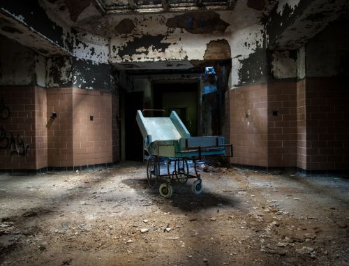 An abandoned, dilapidated room with a rusty cart in the center, surrounded by peeling walls and debris on the floor.