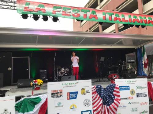 A performer sings on stage at La Festa Italiana, with banners and sponsor logos in the foreground.