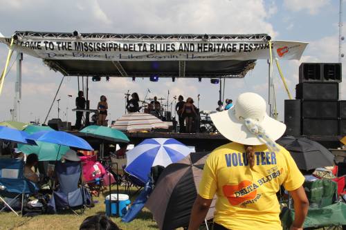 A volunteer watches a band perform on stage at the Mississippi Delta Blues and Heritage Festival, surrounded by umbrellas.