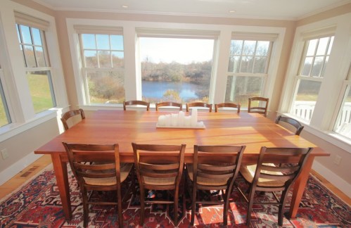 A spacious dining room with a large wooden table and chairs, overlooking a scenic view of a river through large windows.