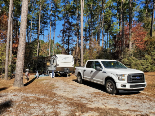 A white pickup truck parked beside a camper in a wooded area with tall trees and autumn foliage.