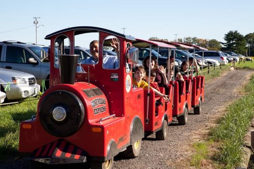 A small red train with children and adults rides along a gravel path, surrounded by parked cars and green fields.