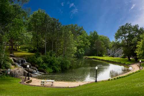 A serene park scene featuring a pond, lush greenery, a small waterfall, and a bench along a walking path.