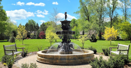 A serene park scene featuring a fountain surrounded by benches and lush greenery under a bright blue sky.