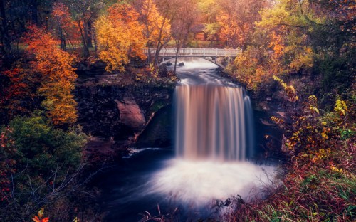 A serene waterfall surrounded by vibrant autumn foliage and a quaint bridge in the background.