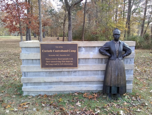 A statue of a woman stands beside a sign marking the site of the Corinth Contraband Camp, surrounded by trees.