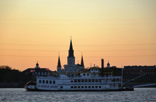 A steamboat named "Creole Queen" sails on water at sunset, with a city skyline featuring church spires in the background.