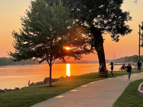 Sunset over a river, with people walking along a path lined by trees and benches, creating a serene atmosphere.