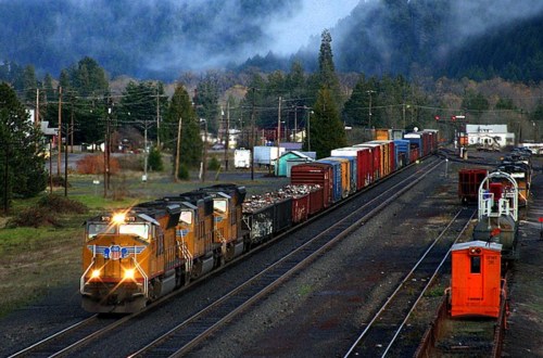 A freight train with colorful cargo cars travels through a scenic landscape with mountains and trees in the background.
