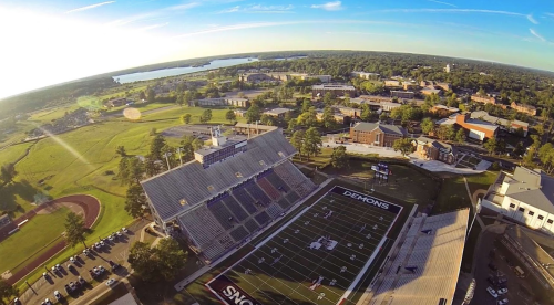 Aerial view of a college campus featuring a football field, surrounding greenery, and buildings under a clear sky.