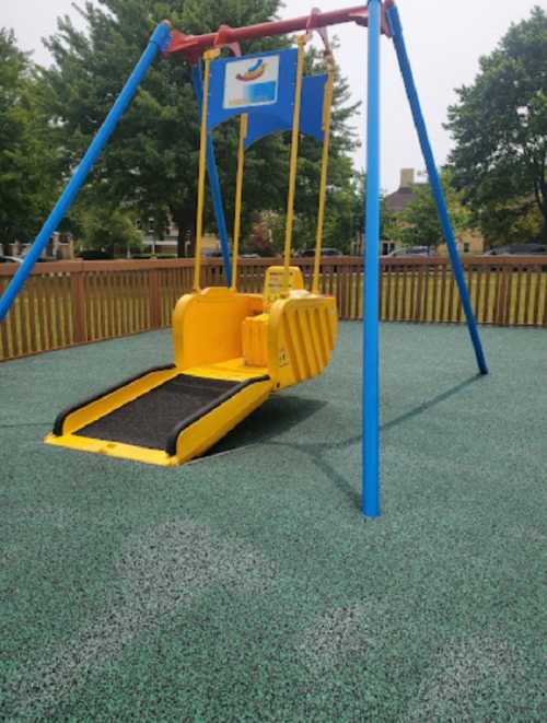 A yellow swing set with a ramp, surrounded by green rubber flooring in a playground. Trees and buildings in the background.