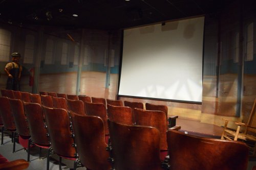 Empty theater with wooden chairs facing a large blank screen and a painted backdrop of a stage.