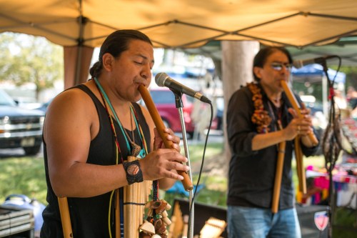 Two musicians play wooden flutes under a tent at an outdoor event, with colorful decorations in the background.