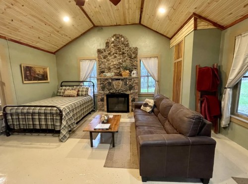 Cozy cabin interior featuring a bed, sofa, stone fireplace, and wooden accents. Natural light from large windows.