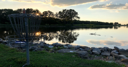 A disc golf basket near a calm lake at sunset, with trees and rocks lining the shore.