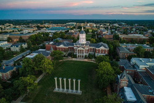 Aerial view of a university campus featuring a central building and four columns on a green lawn at sunset.