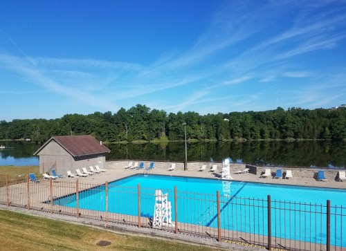A clear blue swimming pool surrounded by lounge chairs, with a grassy area and a calm lake in the background under a blue sky.