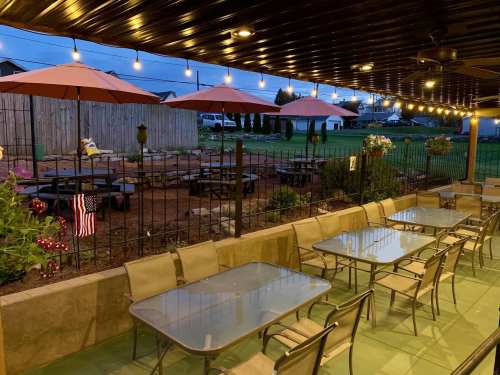 Outdoor dining area with glass tables, umbrellas, and string lights, set against a grassy backdrop at dusk.