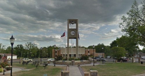 A town square featuring a clock tower, benches, and trees under a cloudy sky.