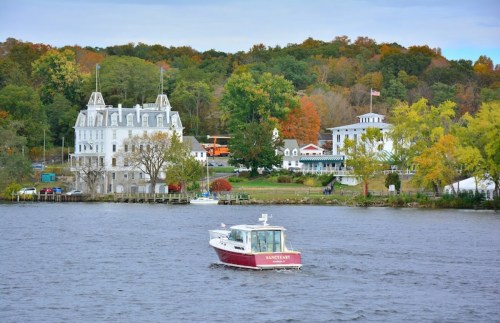 A boat glides on a river near a scenic shoreline with colorful autumn trees and historic buildings.