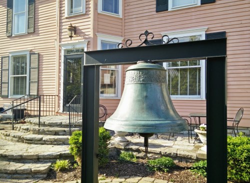 A large bronze bell stands in front of a pink building with black shutters and a stone pathway.