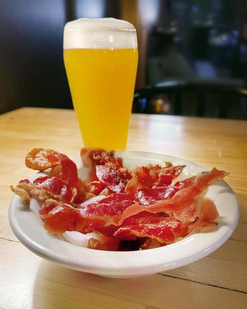 A bowl of crispy bacon strips next to a tall glass of golden beer on a wooden table.
