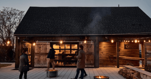 A cozy wooden building with string lights, people walking outside, and smoke rising from fire pits at dusk.