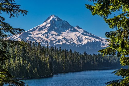 Snow-capped mountain rises above a serene lake, framed by lush green trees under a clear blue sky.
