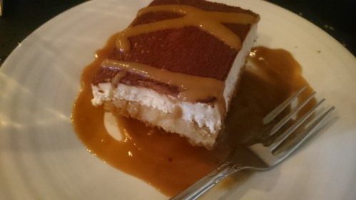A slice of dessert with layers of cream and cake, drizzled with caramel sauce, served on a white plate with a fork.
