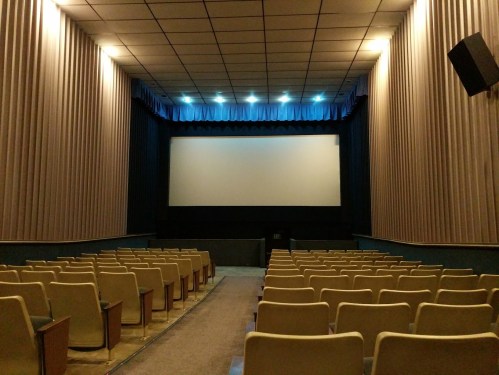 Interior of a small, empty movie theater with rows of beige seats facing a blank screen. Soft lighting illuminates the space.
