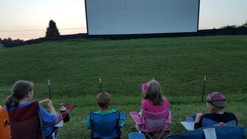 Four children sit in lawn chairs, facing a large outdoor movie screen at dusk, ready for a film.