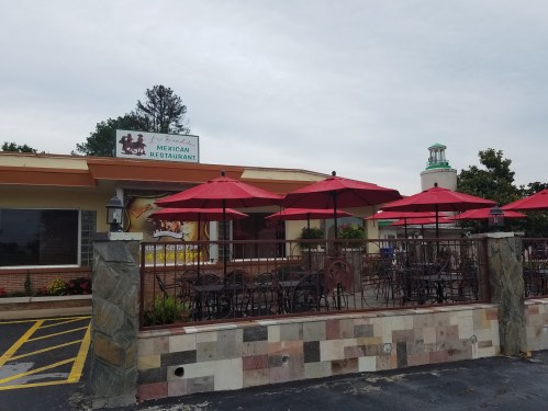 Exterior of a Mexican restaurant with red umbrellas on a patio, featuring a stone wall and a sign above the entrance.