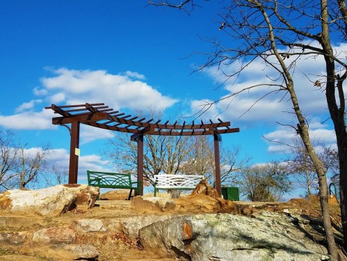 A wooden pergola with two benches sits on rocky terrain under a blue sky with fluffy clouds and bare trees.