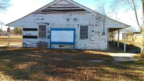 A weathered white building with a blue sign, surrounded by grass and trees, showing signs of age and wear.