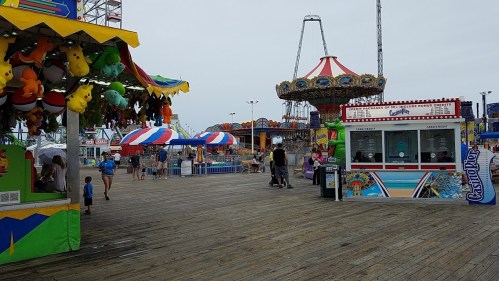 A colorful amusement park scene with games, rides, and people enjoying a cloudy day on a wooden boardwalk.