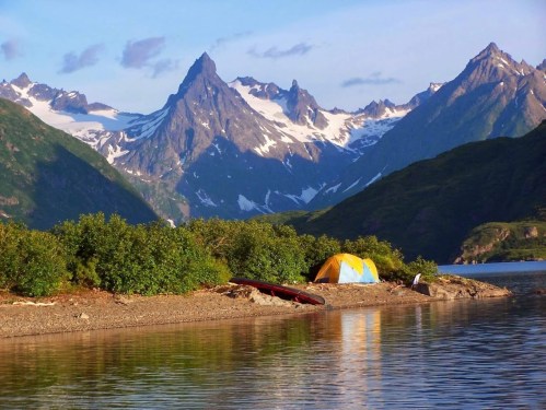 A serene lakeside scene with a yellow tent, surrounded by lush greenery and majestic snow-capped mountains in the background.