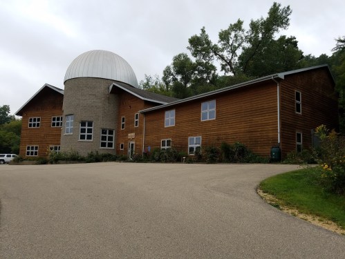 A modern wooden building with a dome, surrounded by greenery and a paved driveway.