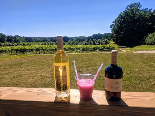 A scenic view of a vineyard with a bottle of white wine, a pink drink, and a bottle of red wine on a wooden railing.