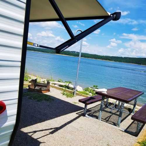 A lakeside view featuring a picnic table, fire pit, and a camper under a shaded awning on a sunny day.