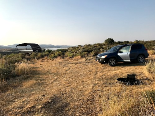 A parked car on a dry, grassy area with a distant view of a lake and a shelter structure under a clear sky.