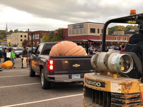 A large pumpkin sits in the bed of a pickup truck at a bustling outdoor event with people and buildings in the background.