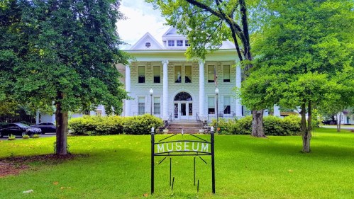 Historic museum building with white exterior, large windows, and a sign in front, surrounded by green trees and grass.