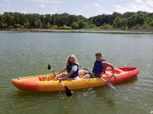 Two people in a kayak on a calm lake, enjoying a sunny day with trees in the background.