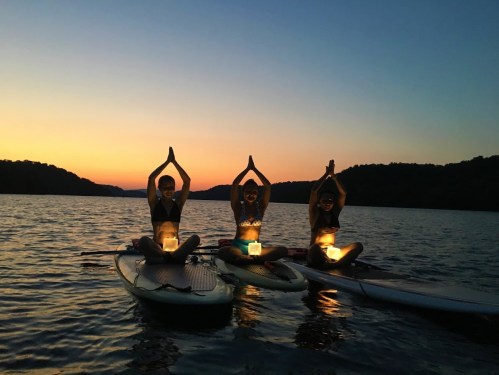 Three people on paddleboards at sunset, practicing yoga with lanterns in front of them, surrounded by calm water.